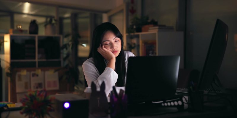 Tired, asian woman and sleeping with laptop at night for deadline, insomnia or fatigue at office. E.