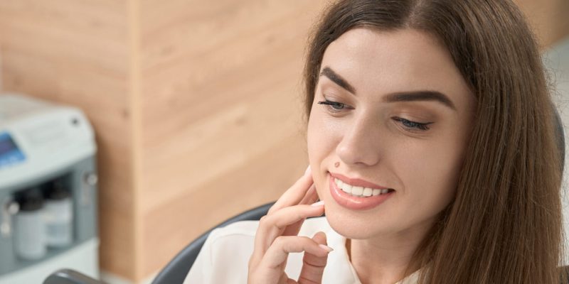 Happy dental clinic customer looking in hand mirror in presence of her stomatologist